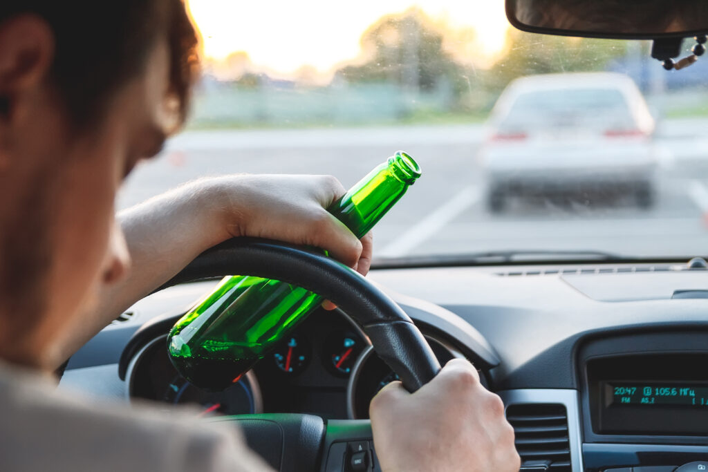 Drunk young man driving a car with a bottle of beer. Distracted Driving