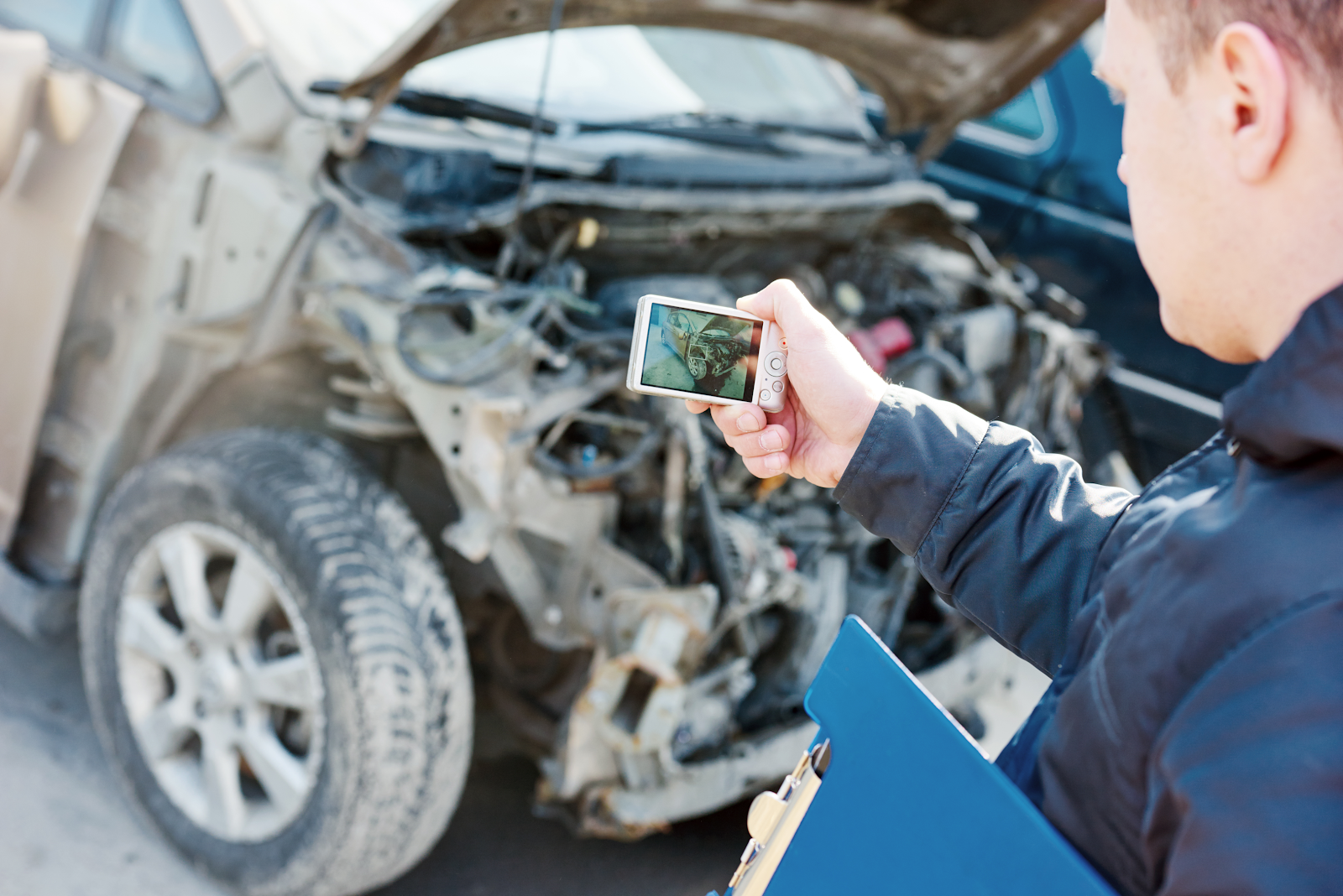 insurance claim adjuster taking a picture of a damaged car with a camera and clipboard in his hand