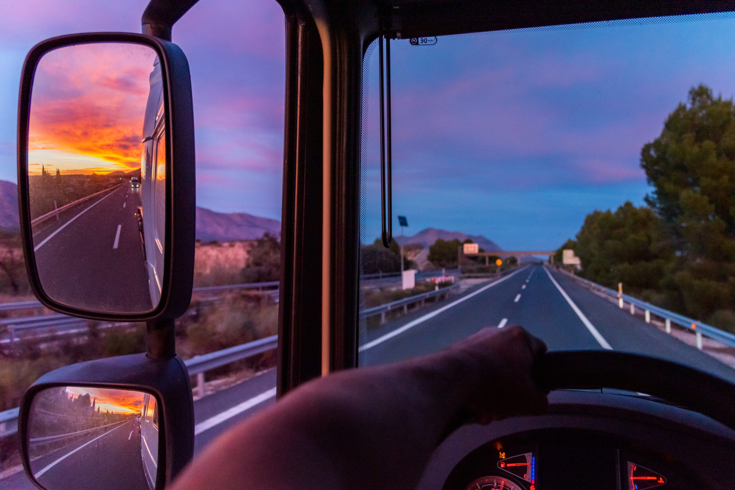 A truck traveling down the highway as the sun sets in the side-view mirror, while cars avoid driving in the trucks blind spots.