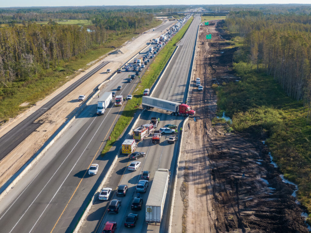 An aerial view of a highway accident involving a large tractor-trailer that has traffic stopped in both directions while first responders are surrounding the crash area.